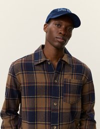 Brązowa i granatowa koszula flanelowa w kratę z zapięciem na guziki, jedną kieszenią na piersi i kontrastowym szwem; połączona z niebieską czapką baseballową.