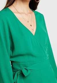 Grüne Wickelbluse aus leichtem Stoff mit tiefem V-Ausschnitt, langen Ärmeln und einem passenden Bindeband in der Taille. Elegante und glatte Textur.