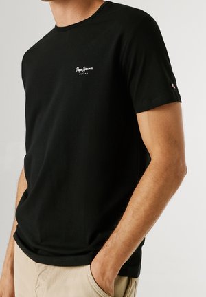Mężczyzna w czarnej koszulce Pepe Jeans London z logo na piersi i beżowych spodniach, z jedną ręką w kieszeni, na jednolitym tle.