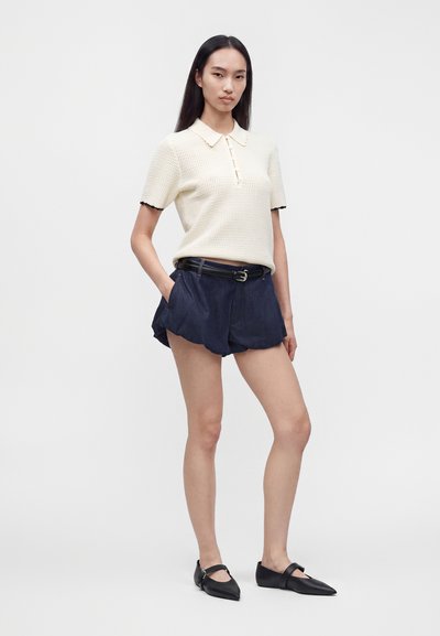 Femme debout les mains dans les poches, portant un polo en maille crème à manches courtes, un short bleu marine avec ceinture et des ballerines noires pointues sur fond blanc.