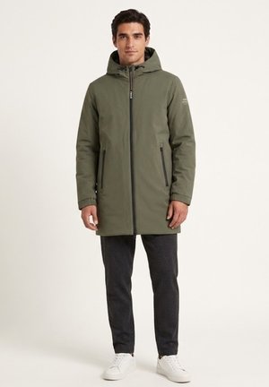Hombre con abrigo de invierno verde oliva con capucha, pantalones oscuros y zapatillas blancas, de pie frente a un fondo claro y liso.
