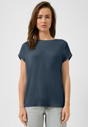 Jeune femme aux longs cheveux bruns, portant un haut ample à manches courtes bleu foncé et un jean gris clair, debout devant un fond uni.