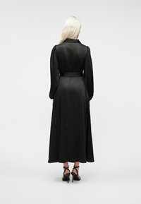 Pikkade blondide juustega naine kannab musta pikakäiselist, vöödiga midikleid ning musti kõrge kontsaga sandaale, seistes valge taustaga.