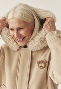Beżowy płaszcz z kapturem wykończonym sztucznym futrem, ozdobiony naszywką w kształcie misia. Tkanina wydaje się miękka, z czystym i uporządkowanym krojem.