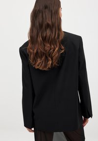 Musta bleiseri, jossa on muotoiltu istuvuus, pitkät hihat, selkeä linja ja sileä pinta. Takakuvassa näkyy vain minimaaliset suunnittelun yksityiskohdat.