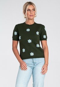 Kobieta ubrana w ciemnozielony sweter z krótkim rękawem w jasnoniebieskie i żółte wzory kwiatowe, w połączeniu z jasnoniebieskimi dżinsami, stoi na białym tle.