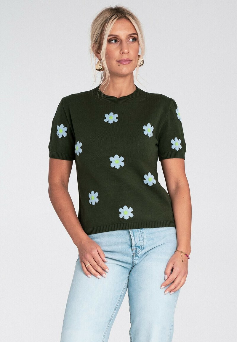 Kobieta ubrana w ciemnozielony sweter z krótkim rękawem w jasnoniebieskie i żółte wzory kwiatowe, w połączeniu z jasnoniebieskimi dżinsami, stoi na białym tle.