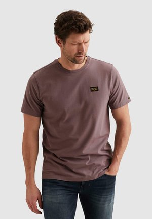 Hombre con camiseta de manga corta color malva con pequeño logo en el pecho, jeans oscuros, mirando hacia abajo con una mano en el bolsillo, sobre un fondo liso.