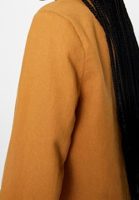 Tissu de couleur moutarde avec une texture lisse, montrant une épaule et une vue de côté taillées, avec de longs cheveux noirs tressés visibles.