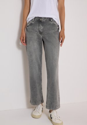 Person iført grå vidde jeans, hvid t-shirt og hvide sneakers med sorte detaljer, der står mod en ensfarvet baggrund.