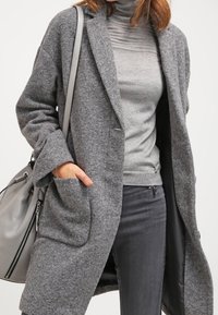 Šedý vlněný kabát s volným střihem, velkými předními kapsami, s klopami, spárovaný se šedým rolákem a tmavě šedými kalhotami. Šedá taška přes rameno.
