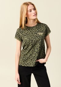 Camiseta verde oliva con un diseño de leopardo negro y mangas cortas enrolladas. Presenta un detalle en texto dorado que dice "reina del baile".