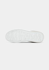 Semelle de sneaker blanche avec un motif texte "GUESS" répété en relief sur le fond contre un arrière-plan blanc uni.