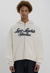 Ung person med krøllet hår bærer en hvid zip-up hættetrøje med teksten "Low Lights Studios" og mørke bukser foran en ensfarvet baggrund.