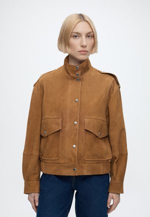 Jeune personne aux cheveux blonds courts portant une veste en daim marron boutonnée avec des poches à rabat et un jean bleu, face à l'avant.