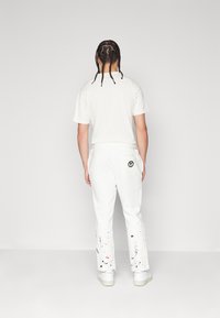 Hvite joggebukser med snor i midjen, med et fargerikt sprutmønster og en logo detalj på baklommen. Kombinert med en hvit t-skjorte.