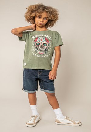 Enfant aux cheveux bouclés portant un t-shirt vert olive avec un motif de crâne coloré, un short en jean, des chaussettes blanches et des baskets beige.