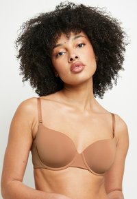 Un reggiseno marrone, imbottito e con stecche, dotato di spalline regolabili. Materiale liscio, coppe senza cuciture e design semplice. Senza decorazioni o fantasie.