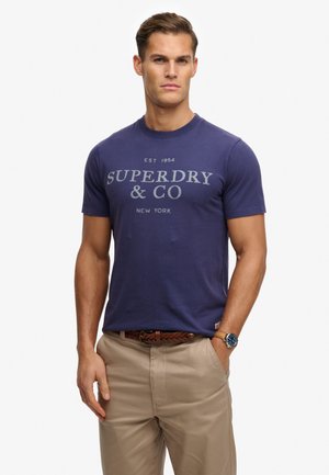 Granatowy bawełniany t-shirt męski z przodu z szarym napisem „SUPERDRY & CO” oraz rokiem założenia, w zestawie z beżowymi spodniami i brązowym plecionym paskiem.