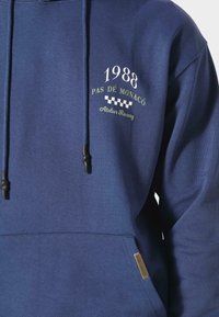 Felpa con cappuccio blu scuro in cotone, con tasca frontale, lacci e testo ricamato: "1988 PAS DE MONACO Atelier Racing."