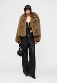 Γυναίκα που φοράει καφέ γούνινο σακάκι, μαύρο τοπ με κοψίματα, μαύρο δερμάτινο παντελόνι φαρδιάς γραμμής, μαύρα μυτερά παπούτσια και κρατάει μια μαύρη τσάντα με στρας.