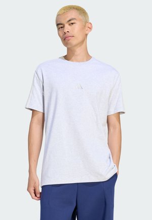 Junger Mann mit kurzen blonden Haaren trägt ein hellgraues Adidas-T-Shirt und blaue Hose, steht mit einer Hand in der Tasche vor hellem Hintergrund.