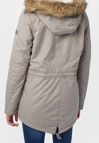 Světle šedá zimní parka s kapucí olemovanou kožešinou, staženým pasem a zipovým ventilačním otvorem na zádech, nošená s modrými džíny.