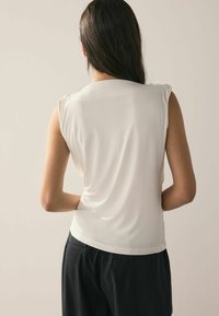 Sleeveless ivory top with a smooth texture and gathered detailing at the shoulders, paired with black trousers featuring side pockets.