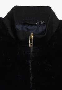 Svart faux fur-jacka med hög krage, dragkedjestängning med guldfärgad detalj och en mjuk innerfoder. Lappen indikerar storlek 8 år.