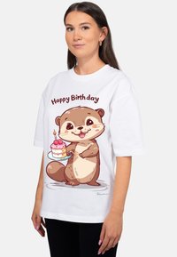 Wit t-shirt met een stripfiguur van een otter die een verjaardagstaart vasthoudt, met de tekst "Happy Birthday" erboven. Korte mouwen en een ontspannen pasvorm.