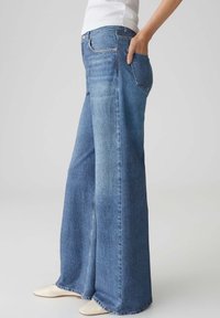 Persoon draagt wijde blauwe spijkerbroek met een hand in de achterzak, gecombineerd met witte schoenen en een wit bovenstuk.