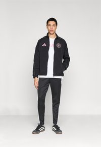 Μαύρο αθλητικό μπουφάν με ροζ λογότυπο και λευκό T-shirt, συνδυασμένο με μαύρο παντελόνι που έχει λευκές λωρίδες στο πλάι και μαύρα αθλητικά παπούτσια.