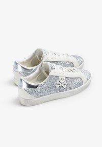 Błyszczące srebrne sneakersy z białymi zamszowymi akcentami, logo czaszki oraz metalicznymi srebrnymi detalami na obcasie i języku. Gumowa podeszwa.