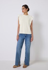 Kvinde med hår i knold iført hvid ærmeløs top med pude skuldre, blå jeans med slids foran og beige sandaler, stående mod en hvid baggrund.