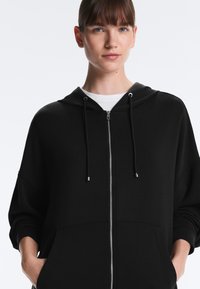 Vrouw met een zwart vest met ritssluiting en handen in de zakken, witte shirt eronder, staand tegen een eenvoudige lichte achtergrond.