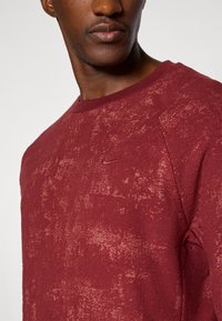 Burgundská mikina s kulatým výstřihem a texturovaným povrchem, s malým logem Nike na hrudi, má pružné manžety a lem.