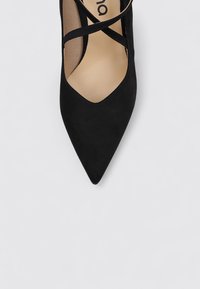 Zapato de tacón negro de ante con detalle de correas entrecruzadas. La textura suave y el diseño minimalista realzan la simplicidad visual.