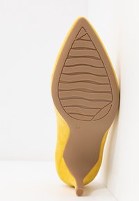 Chaussure à talons hauts jaune avec un bout pointu et une texture en daim. La semelle est en caoutchouc marron, présentant un motif de bande de roulement ondulé et un marquage de taille 37.