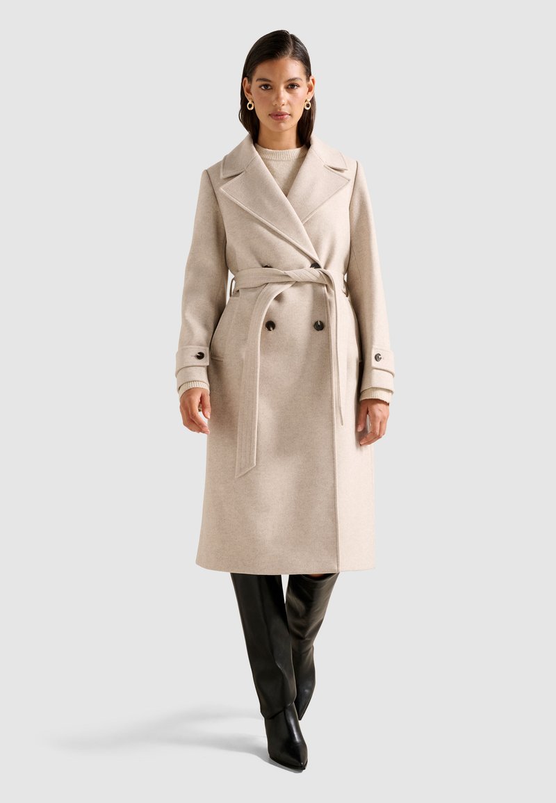 Beige gyapjúkeverék trench coat, övvel a derékon, dupla gombokkal és oldalsó zsebekkel, fekete, testhezálló, térd fölé érő csizmákkal párosítva.