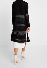 Black crochet cardigan with long sleeves, open knit pattern, and side slits, layered over a white skirt. Features a relaxed fit.