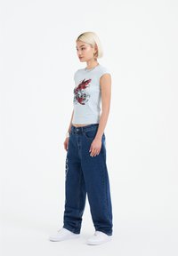 Camiseta ajustada de color azul claro con un gráfico de langosta roja, combinada con unos vaqueros oscuros sueltos que tienen acentos decorativos y zapatillas blancas.