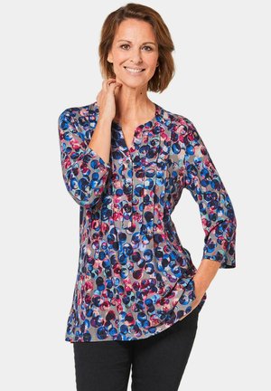 Multikleurige shirt met blauwe, roze en paarse cirkelpatronen, een kraagloze halslijn en driekwart mouwen met plooien.