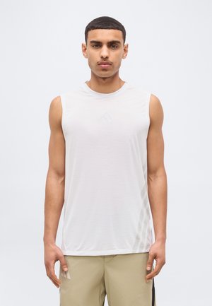 Rövid hajú fiatal férfi, fehér ujjatlan atlétatrikót és bézs rövidnadrágot visel, egyszerű világos háttér előtt áll.