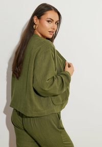 Giacca verde oliva, dal taglio morbido, con tessuto testurizzato, spalle scese e dettagli discreti delle tasche, indossata sopra pantaloni abbinati.