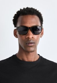 Gafas de sol con montura negra y lentes oscuros. Presentan un acabado glossy con bisagras metálicas, usadas por una persona con una camiseta negra.