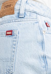 Vaqueros de denim azul claro con una etiqueta de cuero, etiquetas de marca rojas y detalles de botón metálico. Textura ligeramente desgastada en la tela.