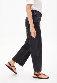 Jeans noirs taille haute avec une coupe droite et des coutures visibles, associés à des sandales plates avec des brides rouges. Modèle vu de profil.