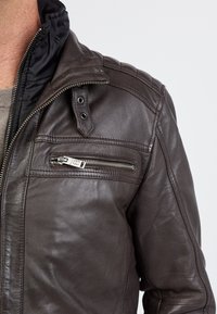 Bruine leren jas met een ritssluiting op de borst en een drukknoopkraag. Heeft gestikte schouderdetails en een gladde, gestructureerde afwerking.