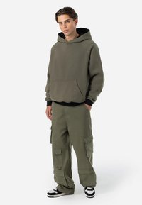 Olijfgroene hoodie met een voorkantzak en zwarte voering, gecombineerd met olijfgroene cargo pants met meerdere zakken en opgerolde cuffs. Zwart-witte sneakers.
