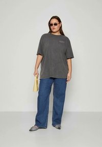 Camiseta gris de gran tamaño con mangas cortas, combinada con jeans de pierna ancha en azul claro. Los accesorios incluyen un pequeño bolso amarillo y zapatillas gris de tipo slip-on.
